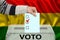 Male voter drops a ballot in a transparent ballot box against the background of the national flag of bolivia, concept of state