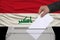 Male voter drops a ballot in a transparent ballot box against the background of the national flag of Iraq, concept of state