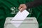 Male voter drops a ballot in a transparent ballot box against the national flag saudi arabia with an Arabic inscription There is