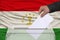 Male voter lowers the ballot in a transparent ballot box against the background of the national flag of Tajikistan, concept of