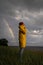 Male in yellow raincoat wear red hat standing on the beach in rainy weather, looks at dramatic cloudy sky and sea, rainbow on