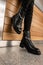 In the mall near a wooden wall stands a young woman in black jeans in trendy leather black autumn lace-up boots. Close-up of