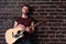 Man with acoustic guitar against brick wall playing music singing songs enjoy life Medium shoot