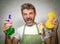 Man in apron holding sponge and detergent spray feeling overwhelmed and bored doing domestic housework of cleaning and washing