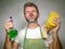 Man in apron holding sponge and detergent spray feeling overwhelmed and bored doing domestic housework of cleaning and washing