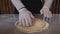 A man in an apron is rolling out pizza dough on a table sprinkled with flour. On the table in front of him are bowls of