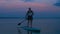 A man with a backpack on a SUP board swims in the lake at dusk in the evening