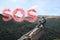 Man with backpack and word SOS made of color smoke bomb on rocky peak