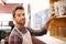 Man, barista and portrait by a shelf in coffee shop with smile for service, search and ingredients for drink. Waiter