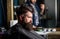Man with beard covered with black cape sits in hairdressers chair, mirror background. Hipster with beard waits for