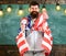 Man with beard on happy face holds flag of USA and clock in classroom, chalkboard on background. American educational