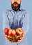 Man with beard holds wicker bowl with fruit on blue background, defocused. Farmer with surprise on face holds red apples