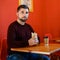 A man with a beard sits in a fast food restaurant and holds a shawarma in his hand and has a glass of coffee.