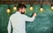 Man with beard stand in front of chalkboard, rear view. Guy busy with writing on chalkboard surface. Bearded hipster in
