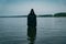 A man in a black robe with a hood stands in the river during the day. He mysteriously looks at the water