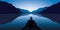 A man on a boat watches a sunrise over a lake in the midst of a sublime mountain landscape.