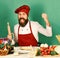 Man with burgundy uniform near table with ingredients on green background. Happy bearded chef cook holding rolling pin