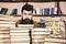 Man on calm face between piles of books, while studying in library, bookshelves on background. Bibliophile concept