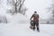 A man in camouflage is walking in the snow and clearing snow with a gasoline-powered snowplow. Back view.