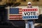 Man with cap, blue jeans and sunglasses holding a cardboard sign text VOTE with american stars and stripes flag