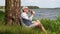 Man in cap sits under a tree on the shore of lake and drinks water