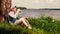 Man in cap sits under a tree on the shore of lake and drinks water