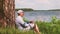 Man in cap sits under a tree on the shore of lake and drinks water