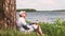 Man in cap sits under a tree on the shore of lake and drinks water