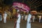 A man carrying a ceremonial umbrella waits as the most sacred elephant at the Kataragama Festival parades through the arena.