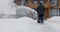 A man cleans the snow with a snowblower near wooden house during a snowfall. Back view. Slow motion