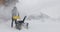 A man cleans the snow with a snowblower near wooden house during a snowfall. Back view. Slow motion