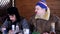 Man in coat sits arond table with food next to his friend with hat ear flaps.