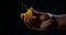 Man is compressing orange slice by fingers, close-up of palm in dark room, juice is squirting in air