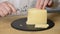 The man cuts cheese into thin slices with use of a cheese slicer on a black round chopping board from slate