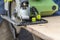 Man cuts a plywood plate using a green electric circular saw tool with yellow chips on a work bench