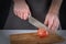 A man in a dark apron slices a tomato into two halves. A beautiful photo of the process of cooking a vegetable , with a