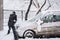 Man in dark clothes cleans his car during heavy snowfall.