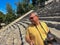 A man in dark glasses and a yellow T-shirt with a camera stands on the steps of the Ancient Theater of Epidaurus. Sanctuary of
