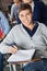 Man With Exam Paper Sitting At Desk In Classroom