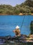 Man fishing with a cane, sitting on rocks with a straw hat on his back.