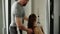 A man fitness instructor in the gym helps a woman use exercise machine and maintains her correct posture. Slender and