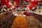 Man in a food stall holding out scoop full of nuts at camera surrounded by vibrant colored fruits and nuts