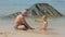 Man and funny son play with sand on ocean beach against rock