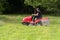 Man gardener driving red modern tractor riding lawn mower in green garden