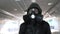 Man in gas mask, black jacket with hood walking through long tunnel, underpass
