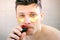 Man with golden patches under eyes shaving moustache using razor looking camera.