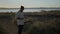 Man guitarist holding a guitar and making a call using a mobile phone while standing on a riverbank at sunset