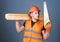 Man, handyman in helmet, hard hat holds handsaw, looks at sharp blade, grey background. Carpenter, woodworker, labourer