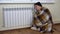 A man in a hat and a plaid at the radiator on the floor. A young man is freezing at home because of non-payment of