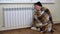 A man in a hat and a plaid at the radiator on the floor. A young man is freezing at home because of non-payment of
